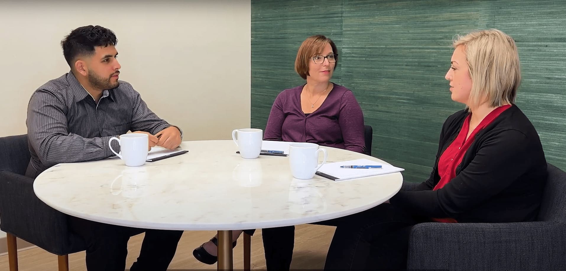 Amy Walls and Sara Bundy from Thimbleberry Financial sitting around a table with a client, looking attentively at each other. There are 3 coffe cups on the table and notebooks with pens.