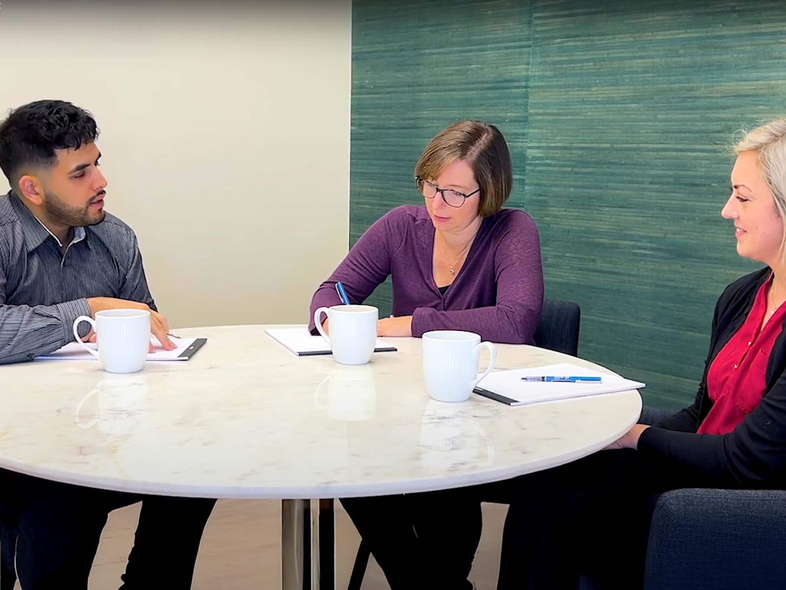 Amy Walls and Sara Bundy from Thimbleberry Financial sitting around a table with a client, looking attentively at each other. There are 3 coffe cups on the table and notebooks with pens. Amy is writing in her notebook